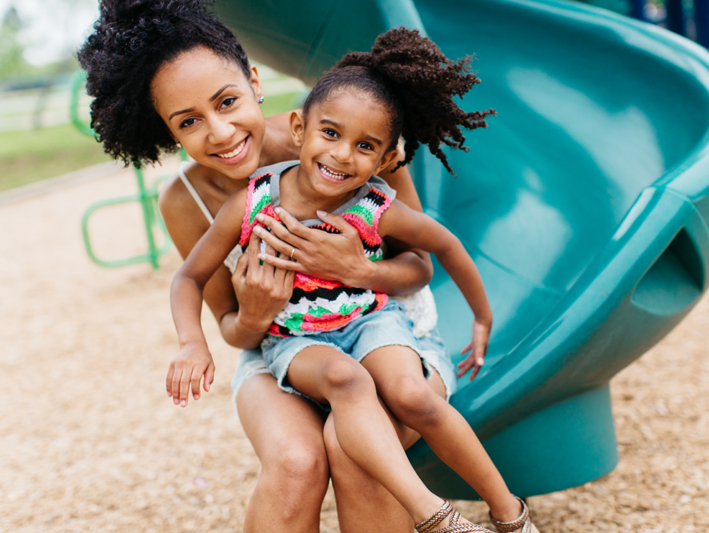An African American mother and daughter are hanging out at the playground. They are posing on a slide and are both smiling. The wind is blowing their hair